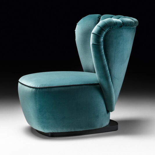 The Designer Upholstered Velvet Occasional Chair is a stylish piece with a unique design featuring a high, rounded backrest that flares outward on both sides. This teal velvet chair has a rounded seat and sits on a black base against a dark gradient background.