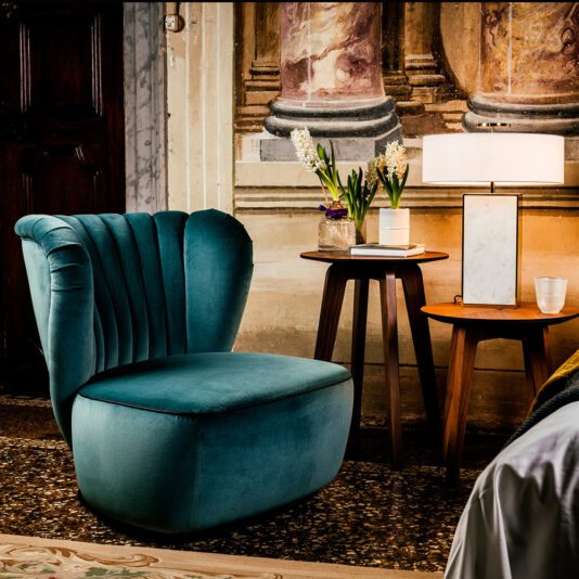 A Designer Upholstered Velvet Occasional Chair sits on a patterned floor beside two wooden tables holding a modern lamp, flowers in vases, and a book. An ornate classical wall mural in the background enhances the room's elegant ambiance.