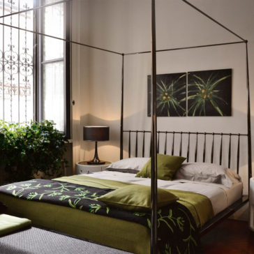 Italian Designer Contemporary Four Poster Bed A cozy bedroom features an **Italian Designer Contemporary Four Poster Bed** adorned with green and black bedding. A potted plant sits near a large window with ornate ironwork. A lamp and two abstract flower paintings add warm accents, creating a serene ambiance.