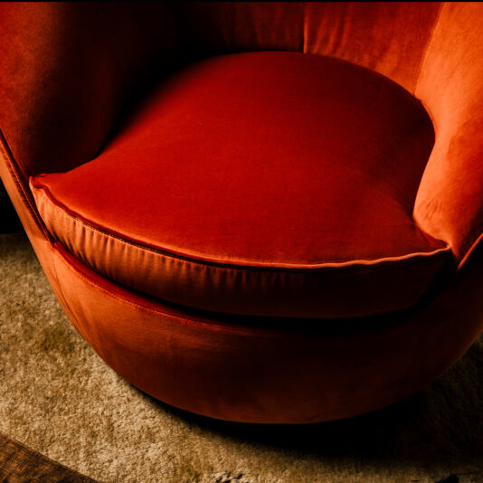 Close-up of the Luxury Designer Velvet Swivel Armchair, featuring a plush, round design in a vivid red-orange velvet with a thick cushioned seat, resting on a muted light brown carpeted floor. The chair's vibrant fabric stands out against the soft, neutral-colored surroundings.