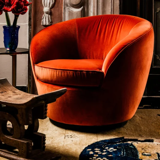 A Luxury Designer Velvet Swivel Armchair in plush orange with a rounded back rests on a patterned rug next to a small table topped by a blue vase with red flowers, all set in an ornate, warmly lit room.