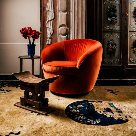 A cozy space features a Luxury Designer Velvet Swivel Armchair in vibrant orange beside a small table holding a blue vase of red flowers. A decorative wooden stool rests on an artfully patterned rug, with ornate doors creating an elegant backdrop.