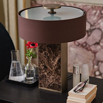 Modern Marble Designer Bronze Table Lamp A stylish bedside table setup features a Modern Marble Designer Bronze Table Lamp with a round brown lampshade. Below the lamp is a black tray holding a glass carafe and a small vase with a red flower. Nearby are books, a pair of glasses, and a lotion bottle, completing the chic arrangement.