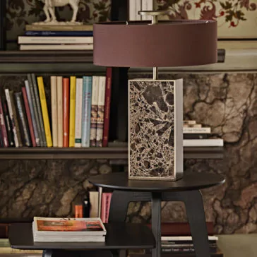 Modern Marble Designer Bronze Table Lamp A Modern Marble Designer Bronze Table Lamp rests elegantly on a dark wood table. Behind the lamp, bookshelves are filled with colorful books. Nearby, a smaller table holds an open magazine, while in the background, there is a wall featuring a floral-patterned section.