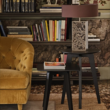Modern Marble Designer Bronze Table Lamp A cozy living room featuring a mustard velvet tufted sofa next to a set of modern black nesting tables. The larger table holds a unique Modern Marble Designer Bronze Table Lamp, while the smaller table has a book on top. A bookshelf with colorful books and decor is in the background.