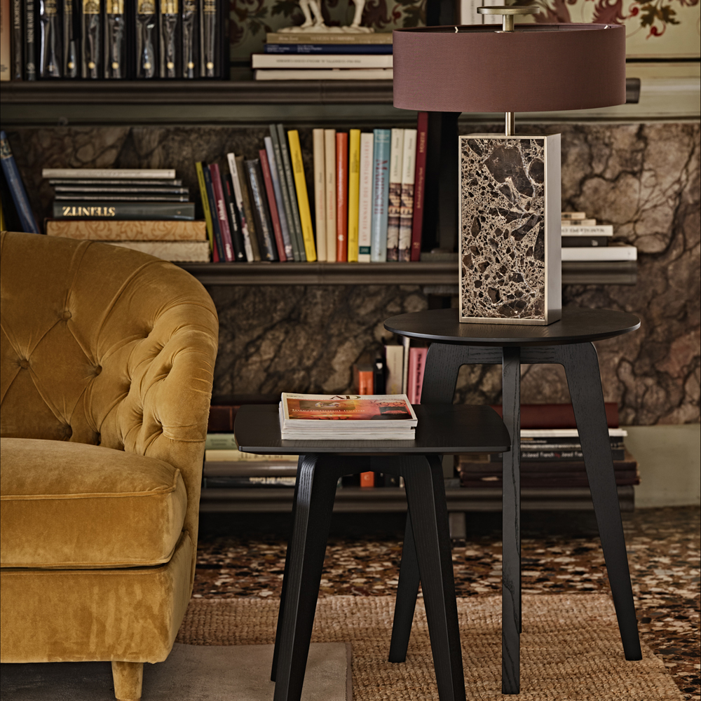 A cozy living room featuring a mustard velvet tufted sofa next to a set of modern black nesting tables. The larger table holds a unique Modern Marble Designer Bronze Table Lamp, while the smaller table has a book on top. A bookshelf with colorful books and decor is in the background.