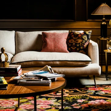 Contemporary Designer Modular Linen Sofa In a cozy living room, the Contemporary Designer Modular Linen Sofa is adorned with two pillows, one red and one floral. A wooden coffee table hosts books and magazines, set atop a colorful floral rug. Warm light from a table lamp completes the inviting atmosphere.