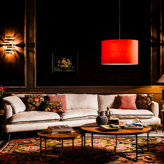 A cozy living room featuring the Contemporary Designer Modular Linen Sofa adorned with colorful patterned pillows, a vibrant red pendant lamp overhead, and two wooden coffee tables topped with decorative items. The dark walls showcase framed artwork, while a patterned rug adds warmth to the space.