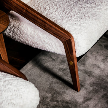 Close-up of the Designer Contemporary Furry Walnut Arm Chair with plush white faux fur upholstery and walnut wooden armrests. The chair's leg is elegantly curved, showcasing detailed craftsmanship. The furry chair is set on a gray carpet, perfectly paired with a matching footstool for added comfort.