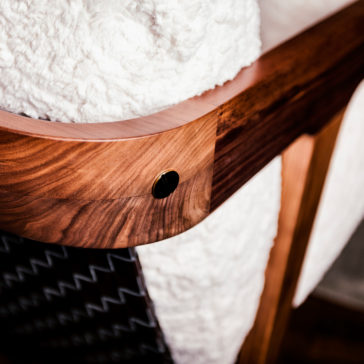 Close-up image of a Designer Contemporary Furry Walnut Arm Chair corner with a dark button and a soft, white textured blanket draped over the chair. The walnut wood has a smooth finish and displays natural grain patterns. The soft blanket contrasts with the wood, conveying a cozy and warm atmosphere reminiscent of designer furniture.