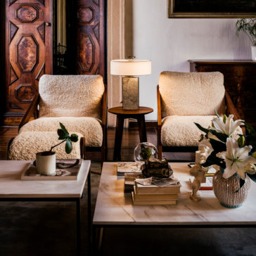 A cozy living room featuring two Designer Contemporary Furry Walnut Arm Chairs with plush white cushions and matching footstools. Between them is a small table with a white marble lamp. In front, there are two contemporary coffee tables adorned with books, a potted plant, and a vase of white lilies. A walnut cabinet completes the background scene.