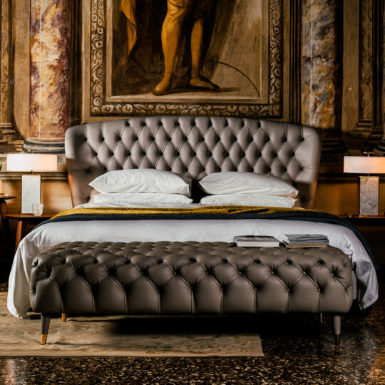 A luxurious bedroom showcasing the Designer Leather Button Upholstered Luxury Bed with a matching bench, white bedding, and bedside tables. Ornate columns and a classical painting create an elegant, opulent ambiance.