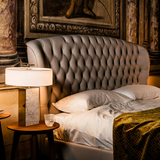 Designer Leather Button Upholstered Luxury Bed A luxurious bedroom showcases the Designer Leather Button Upholstered Luxury Bed, accented with pristine white pillows, a shimmering gold quilt, and a sleek modern table lamp resting on a wooden bedside table, all set against an ornate classical-style mural wall.