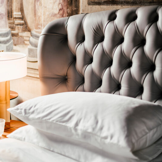 Designer Leather Button Upholstered Luxury Bed Close-up of a bedroom showcasing the Designer Leather Button Upholstered Luxury Bed, featuring a tufted headboard in soft grey leather, white pillows, and part of a bedside table with a modern lamp. The background has an elegant mural with architectural elements, giving the room a designer ambiance.