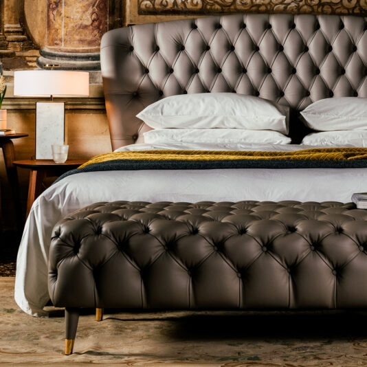 Designer Leather Button Upholstered Luxury Bed Luxurious bedroom featuring the Designer Leather Button Upholstered Luxury Bed and a matching bench in front of a neatly made bed with white linens and blue accents. A wooden bedside table with a lamp, books, and a plant adds warmth to the elegant setting. Ornate wall details are visible in the background.