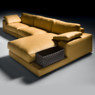 Exclusive Linen Modular Chaise Style Corner Sofa Presenting the Exclusive Linen Modular Chaise Style Corner Sofa, a modern L-shaped sectional in mustard yellow. It boasts thick cushions and a sleek design, with an armrest that holds a woven basket and a pillow perched on the backrest. Set against a flat, dark surface with minimal background distraction, this sofa is both stylish and functional.