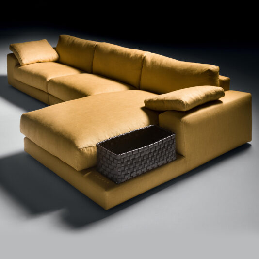 Exclusive Linen Modular Chaise Style Corner Sofa Presenting the Exclusive Linen Modular Chaise Style Corner Sofa, a modern L-shaped sectional in mustard yellow. It boasts thick cushions and a sleek design, with an armrest that holds a woven basket and a pillow perched on the backrest. Set against a flat, dark surface with minimal background distraction, this sofa is both stylish and functional.