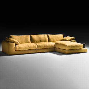 Exclusive Linen Modular Chaise Style Corner Sofa An Exclusive Linen Modular Chaise Style Corner Sofa, adorned in a modern L-shaped design with a striking mustard yellow hue, stands out against a dark backdrop. The sofa boasts wide cushions, a chaise lounge on one end, and two large armrest cushions flanking both sides. It's set upon a light grey floor.