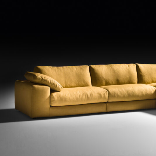 Exclusive Linen Modular Chaise Style Corner Sofa An Exclusive Linen Modular Chaise Style Corner Sofa in a modern mustard yellow is centered in the image against a black background. The exclusive linen cushions and low backrest enhance its contemporary look. The left armrest is visible, and one of the back cushions is slightly angled, adding to its modular charm.