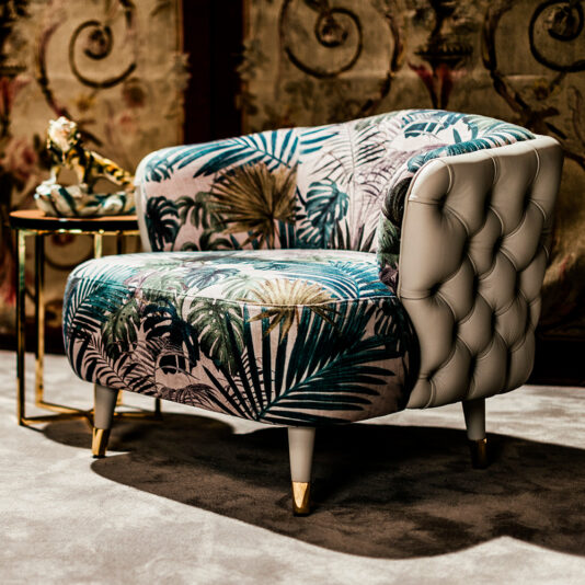A Leather Button Upholstered Designer Botanical Print Tub Chair, featuring a tufted leather back, graces a plush carpet. Its wooden legs accented with gold tips enhance its sophisticated aesthetic, while a small gold side table nearby holds an elegant decorative sculpture.