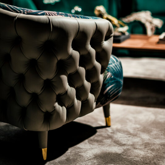 Leather Button Upholstered Designer Botanical Print Tub Chair A close-up reveals the plush texture of the Leather Button Upholstered Designer Botanical Print Tub Chair, complete with its luxurious golden-tipped legs. The chair's rich texture and opulent design are beautifully accentuated by soft, ambient lighting. In the background, a coffee table adorned with a decorative item remains blurred.