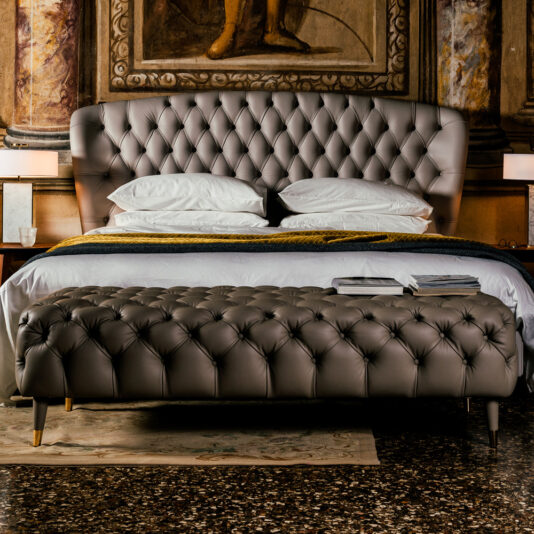 An elegant bedroom showcases a plush, tufted gray leather bed with crisp white pillows and a dark blanket. At the foot stands the Designer Button Upholstered Bench. The ambiance is completed by ornate wall art and a glossy stone floor.