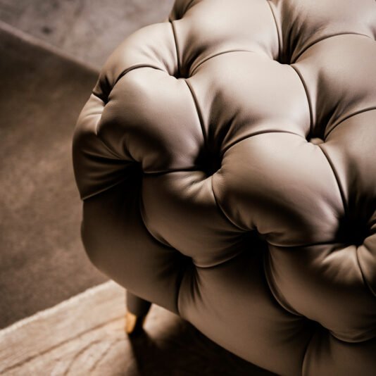 Close-up view of the luxurious Leather Designer Button Upholstered Bench in tan leather, featuring deep button details. Positioned on a two-toned floor of carpet and hardwood, the upholstered bench's soft folds and plush texture are accentuated by the warm, ambient lighting.