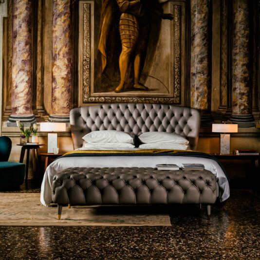 Designer Leather Button Upholstered Luxury Bed A lavish bedroom features a Designer Leather Button Upholstered Luxury Bed with bench, an opulent mural with a historical figure, elegant lamps, a green armchair, and a rug on the dark patterned floor, creating a sophisticated ambiance.