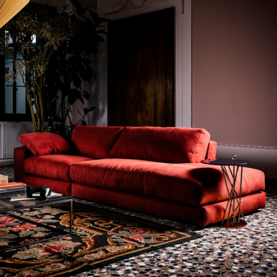 A Luxury Linen Modular Chaise Style Sofa sits in a sophisticated living room with a patterned rug, a sleek glass coffee table, and a small round side table. The room features a mix of contemporary and vintage elements, including a large plant in the background and a wooden door.