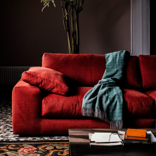 A Luxury Linen Modular Chaise Style Sofa, adorned with a matching cushion and a teal patterned throw, is positioned in a dimly lit room. A coffee table in the foreground holds a stack of notebooks and some loose papers. The floor features a patterned rug, and a potted plant is in the background.