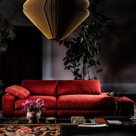 A stylish living room features a large Luxury Linen Modular Chaise Style Sofa with a single cushion, adorned with a pink flower arrangement in a vase on the rectangular wooden table in front. A large gold pendant light hangs above, and a tall plant adds greenery to the corner.