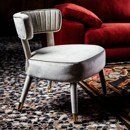 A Modern Designer Velvet Occasional Armchair, upholstered in light gray fabric with gold-tipped legs, stands on a patterned floor. This chair features a rounded seat and a semi-circular backrest with vertical stitching. A red sofa with a pillow is visible in the background.