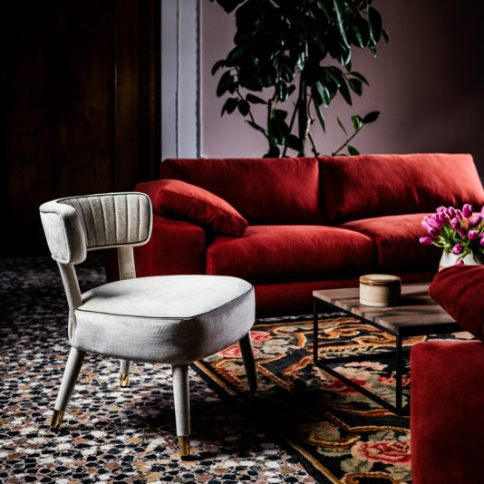 A stylish living room with a white upholstered chair, a red sofa, and a coffee table adorned with a vase of pink flowers. A potted plant stands in the background. The floor features a mosaic pattern and a floral rug. The Modern Designer Velvet Occasional Armchair adds an elegant touch to the softly painted walls.