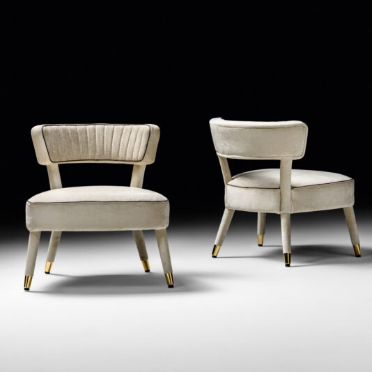 Displayed against a black and white gradient background, two elegant Modern Designer Velvet Occasional Armchairs with beige upholstery and gold-tipped legs emanate sophistication. One chair is shown from the front, spotlighting its ribbed backrest, while the other is slightly turned to highlight its curved design, perfect for use as an occasional armchair.