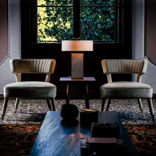 A cozy room featuring two Modern Designer Velvet Occasional Armchairs with wooden legs, positioned on either side of a small wooden table with a marble lamp on top. A large window with closed brown shutters and a view of green foliage is in the background. A patterned rug covers the floor.
