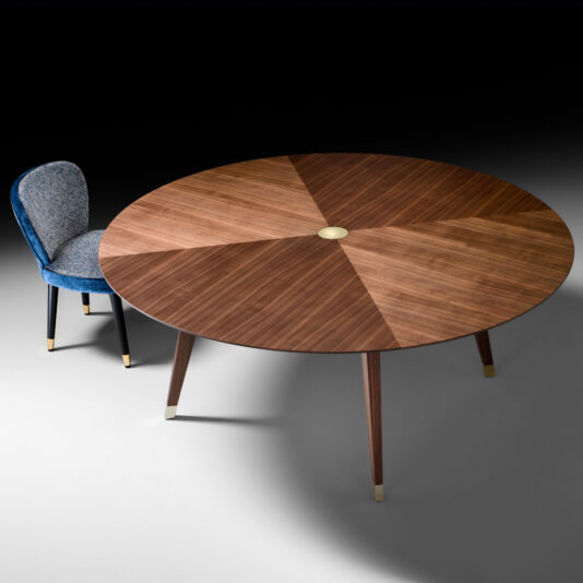 A Contemporary Round Walnut Designer Dining Table with a segmented design and metal-capped legs stands next to an upholstered chair with a rounded backrest and metal tips on the legs. The chair, featuring blue fabric and a black base, creates a stylish, modern contrast to the table's wood grain pattern.