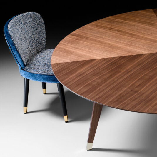 Contemporary Round Walnut Designer Dining Table A stylish **Contemporary Round Walnut Designer Dining Table** with a minimalist design sits next to a plush blue chair with a patterned backrest. Both the table and chair legs feature metallic gold accents at the tips, adding a touch of elegance to this furniture set.