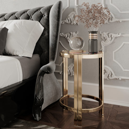 A luxurious Art Deco-inspired bedroom with a gray upholstered bed, white and gray bedding, and an Art Deco Inspired Designer Contemporary Luxury Side Table. The table holds a glass orb and a cylindrical vase with dried flowers. The room features intricate wall paneling and a herringbone wooden floor.