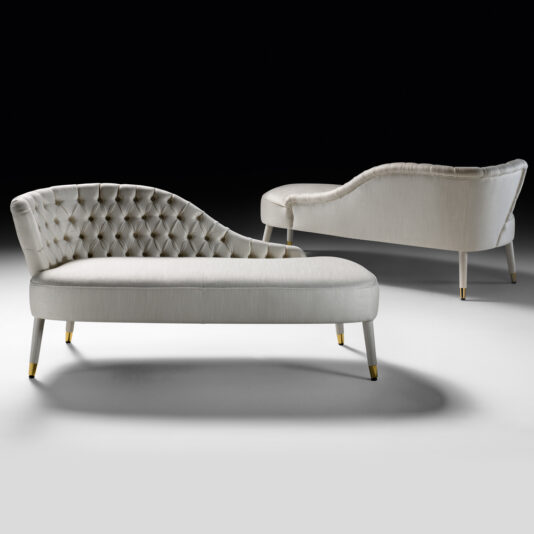 Button Upholstered Designer Linen Chaise Longue Two Button Upholstered Designer Linen Chaise Longues, featuring curved backrests and smooth linen upholstery in a light color, are set against a black background. The chaise longues showcase sleek, tapered legs with gold-toned accents at the feet.