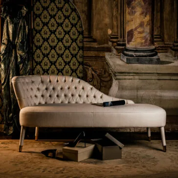 The Button Upholstered Designer Linen Chaise Longue with tufted detailing sits in an ornate room. On its seat rests a book, while black high heels and shoeboxes are placed on the rug amid antique decor and patterned wallpaper.