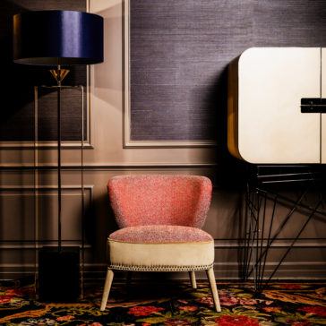 Contemporary Two-Tone Upholstered Designer Occasional Chair A stylish interior featuring a Contemporary Two-Tone Upholstered Designer Occasional Chair with nailhead trim. To the left, a modern floor lamp with a blue lampshade and transparent base stands on a carpet with a bright floral pattern. A beige cabinet with black accents is on the right.