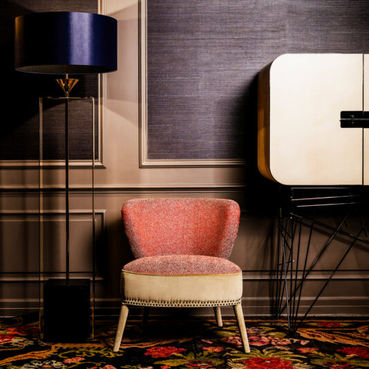 A stylish interior featuring a Contemporary Two-Tone Upholstered Designer Occasional Chair with nailhead trim. To the left, a modern floor lamp with a blue lampshade and transparent base stands on a carpet with a bright floral pattern. A beige cabinet with black accents is on the right.
