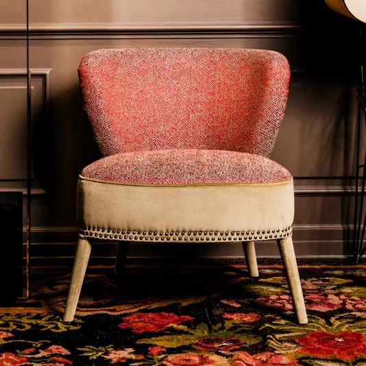 The Contemporary Two-Tone Upholstered Designer Occasional Chair features red patterned backrest and seat cushions, a beige lower section with nailhead trim, wooden legs, and is placed on a floral rug against a paneled wall.