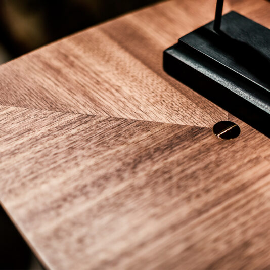 Close-up of a polished walnut surface featuring a geometric pattern with intersecting lines. A small, round, brass-colored screw is embedded neatly in the wood. The base of a black metal object, part of the Contemporary Walnut Designer Console Table, is seen at the top right corner of the image.