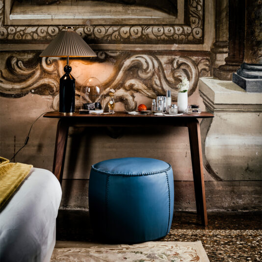 A Contemporary Walnut Designer Console Table, adorned with a black lamp, toiletries, and various small items, stands against a mural wall. A blue cushioned stool is positioned in front of the table. Part of a bed with a yellow blanket is visible on the left, and the floor features a patterned rug.