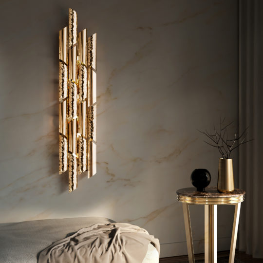 A modern, minimalist room features intricate wall art with gold accents. Below, a round gold table holds a black vase with bare branches and another black decorative item. Adding an extra touch of elegance above the bed with light gray bedding on the left is the Luxurious 24 Carat Gold Plated Contemporary Wall Light.