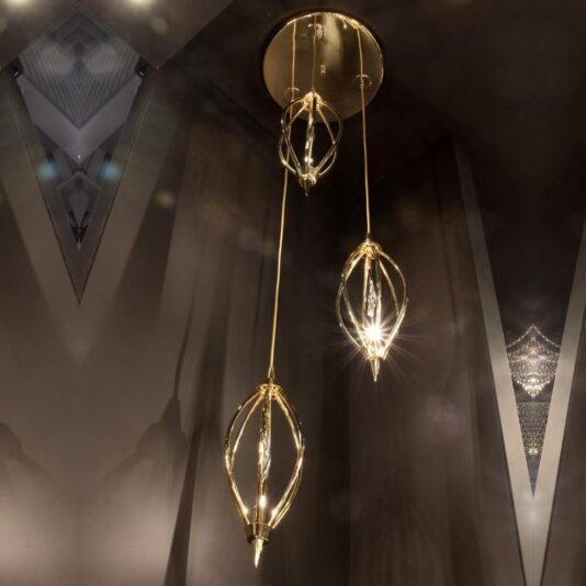 The Modern Crystal Triple Pendant Chandelier features three elongated lights suspended from a circular ceiling mount. Its gold fixtures boast a geometric, cage-like design, casting warm, ambient light that beautifully illuminates a dimly-lit room with dark walls.