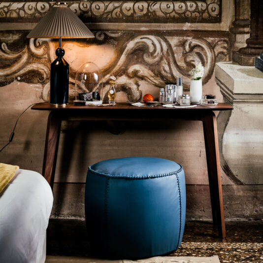 A Contemporary Walnut Designer Console Table creates a chic bedside arrangement, showcasing a lamp with a pleated shade, two drinking glasses, a bowl containing an orange, and several small cosmetic containers. Positioned beneath the table is a blue upholstered ottoman, all set against an ornate fresco-style wall decoration.