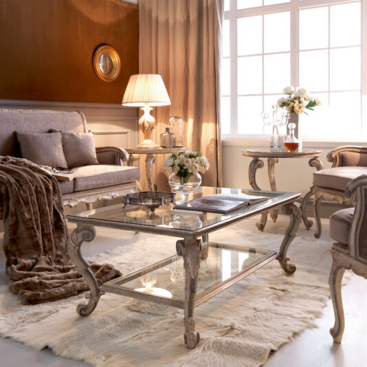 Classic Italian Ornate Designer Glass Coffee Table A luxurious living room with vintage-style furniture, including a Classic Italian Ornate Designer Glass Coffee Table, plush armchairs, and a sofa draped with a fur throw. A designer table lamp with a beige shade sits on a side table. Large windows with light curtains allow natural light to fill the room.
