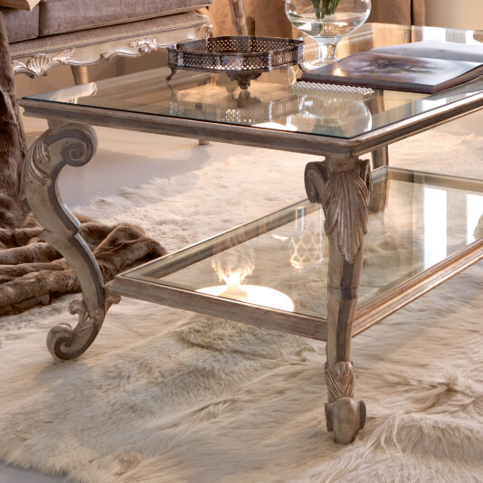 Classic Italian Ornate Designer Glass Coffee Table An elegant living room scene showcases a light, fluffy rug adorned with a Classic Italian Ornate Designer Glass Coffee Table. The intricately carved details on the table's legs and lower border are highlighted, enhancing its sophisticated appeal. Completing the look, a decorative tray and an open book rest gracefully atop the table.